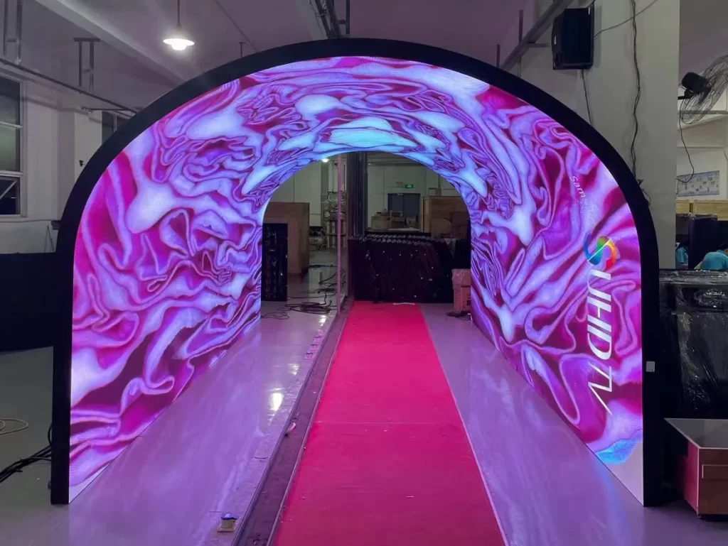 A vivid LED display tunnel with a U-shaped arch, showcasing swirling pink and purple visuals across its flexible digital panels. A red carpet runs through the center, inviting attendees into an immersive, futuristic experience. The tunnel is part of a trade show setup, designed to captivate and engage visitors.